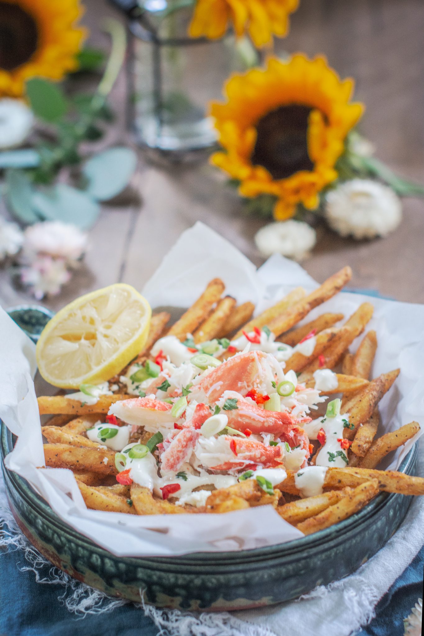 Loaded Crab Fries ruskealla pöydällä, taustalla auringonkukkia